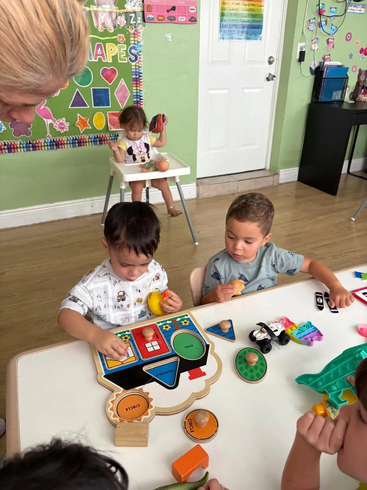 Children sit at a table playing with shape puzzles and toys while a toddler in a high chair holds a toy and an adult's head is visible in the foreground.