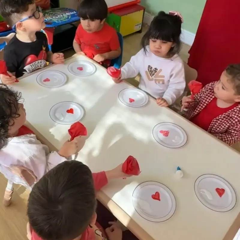 Six children sit around a white table with paper plates and red tissue paper hearts, holding red craft materials.
