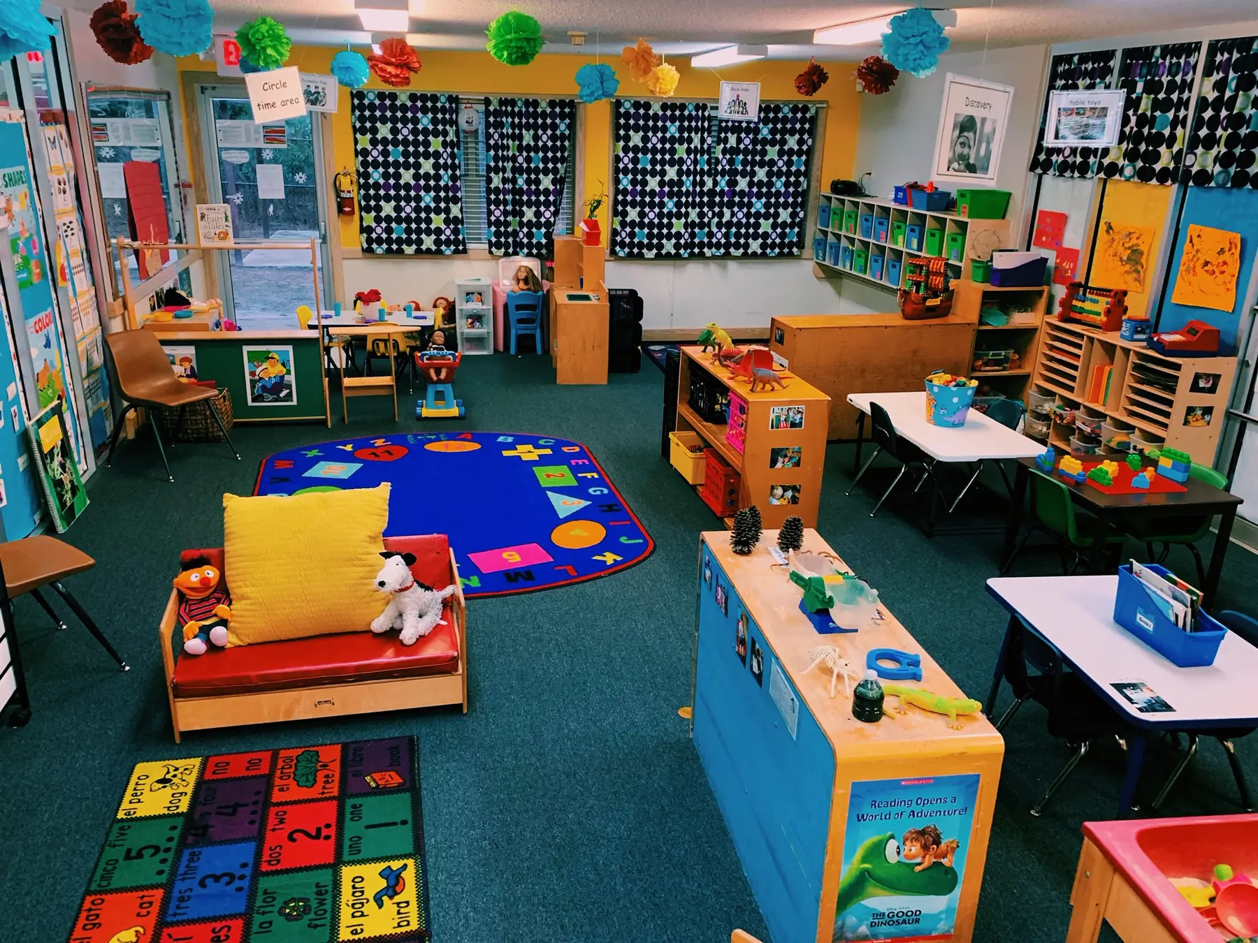 A brightly decorated childcare classroom with colorful rugs, toy shelves, a red couch with stuffed animals, and learning materials arranged around the room.