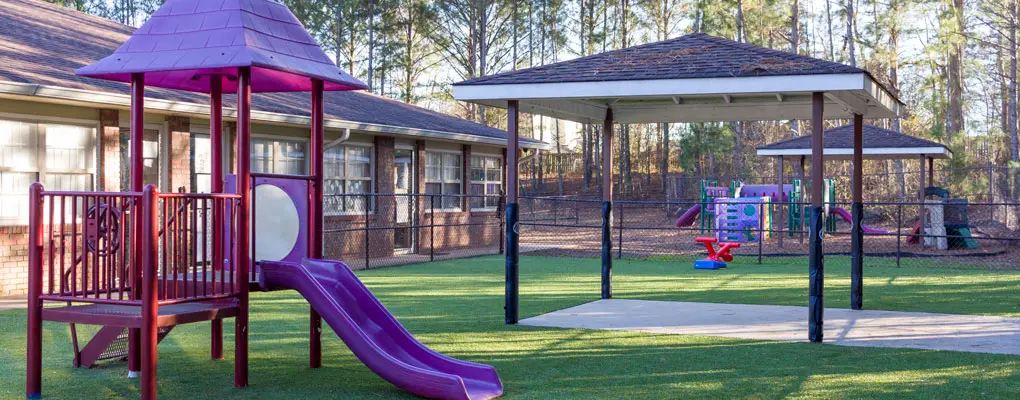 A fenced outdoor play area with a purple slide structure, a covered pavilion, and a small playhouse sits beside a building and tall pine trees.