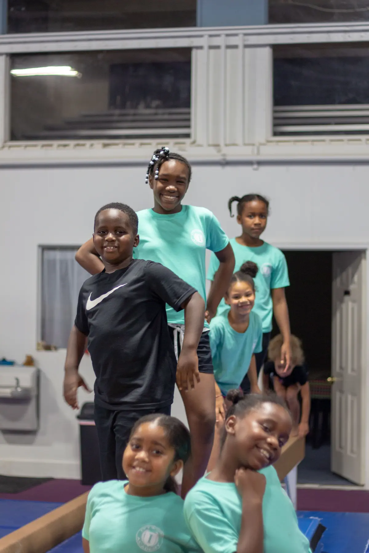 Six children stand and sit on and around a wooden balance beam in a gym, with one boy in a black shirt and five girls in matching teal shirts smiling at the camera.