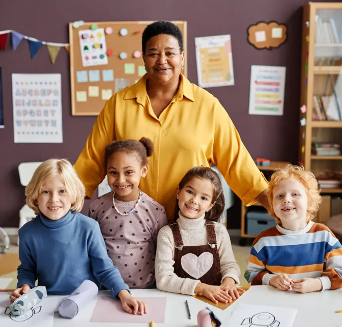 An adult in a yellow shirt stands behind four children seated at a table with paper and craft supplies in a classroom.