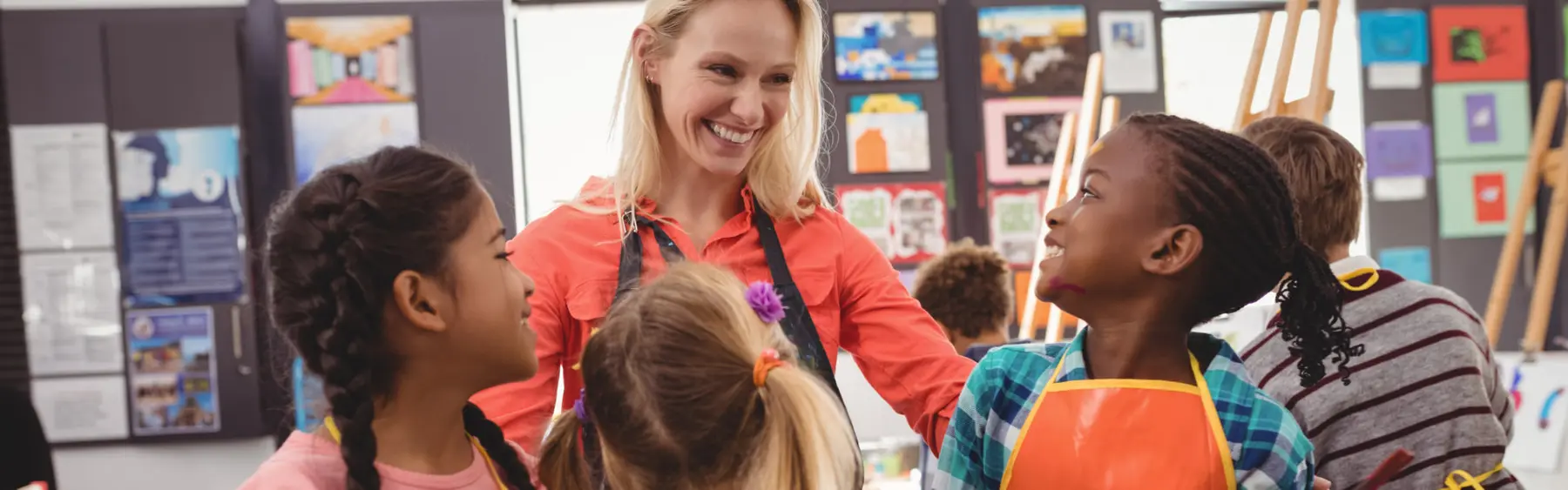 A smiling adult in an orange shirt and black apron stands among several children wearing art smocks in a room with artwork and easels on the walls.