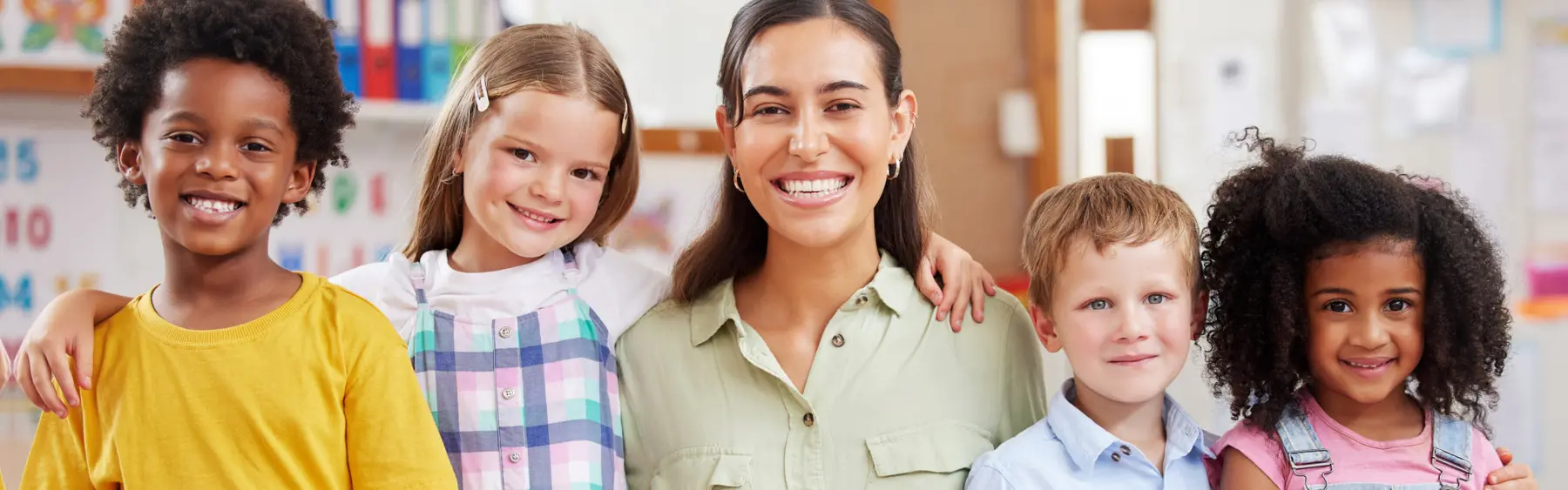 A woman stands in the center with four children beside her, all facing the camera and smiling in front of a classroom background.
