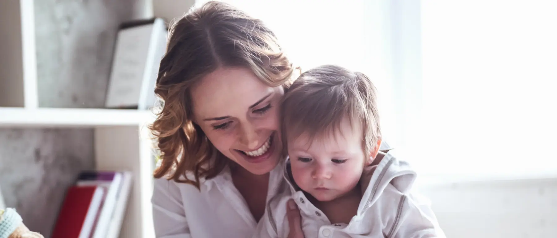 A smiling woman holds a baby in a white hooded top near a bright window, with a shelf of books behind them.