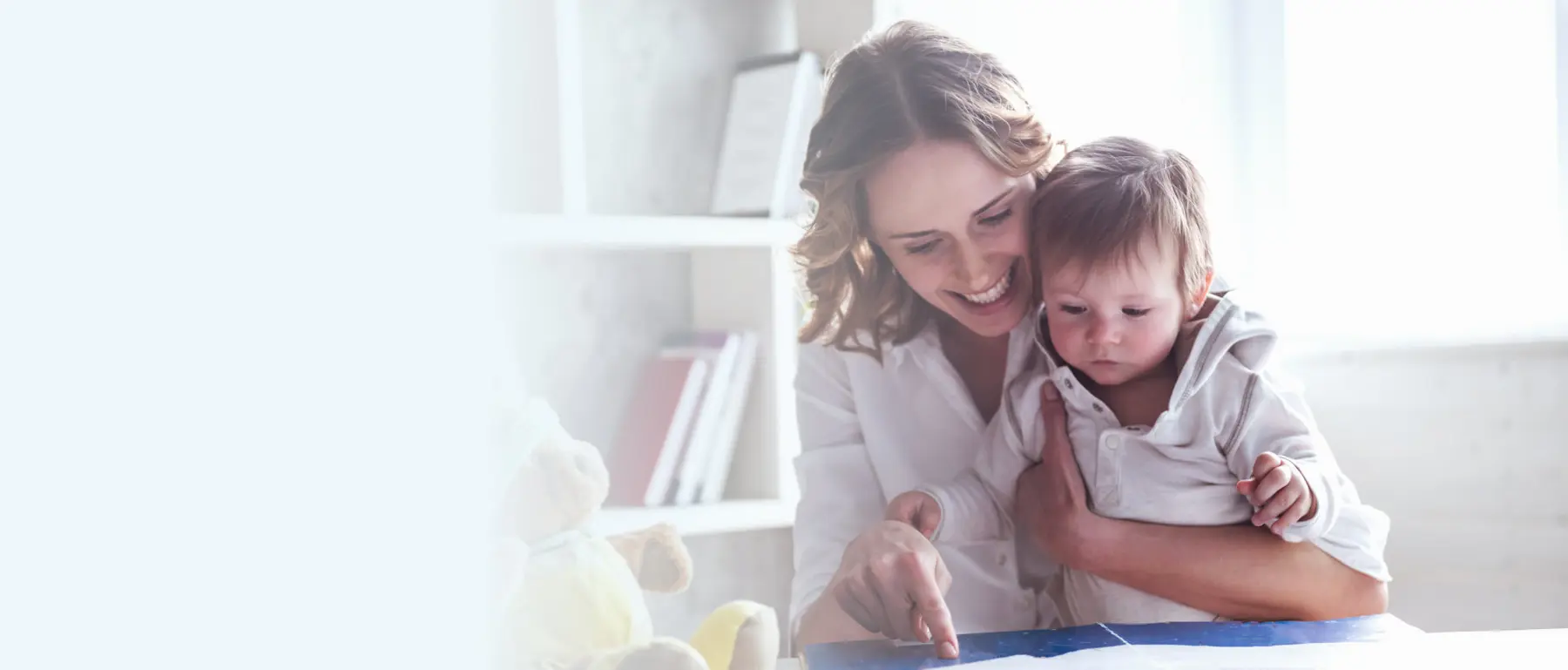 A smiling woman holds a baby on her lap and points at a blue book or tablet on a table, with a white teddy bear and a shelf of books behind them.
