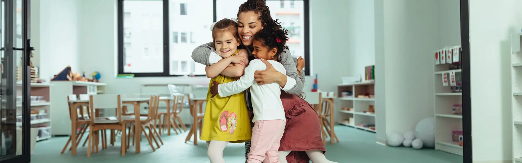 A woman kneels on a green floor and hugs two young girls in a room with wooden tables, chairs, and shelves.
