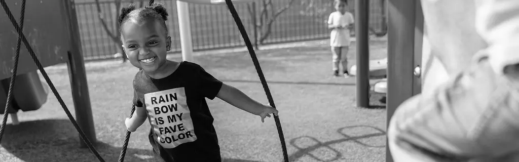 A smiling girl in a black shirt with white text holds playground ropes while another child stands near a fence in the background.