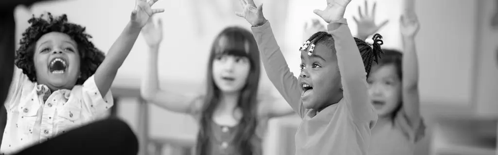 Four children raise their hands with excited expressions in a classroom setting.