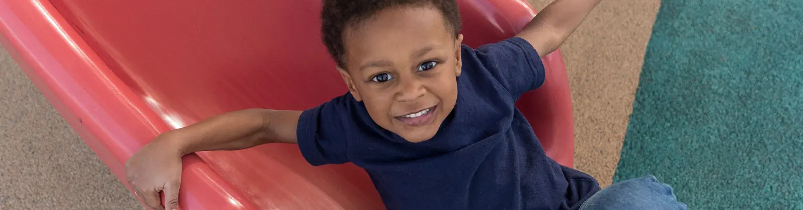 A smiling boy in a blue shirt sits on a red plastic slide with his arms outstretched.