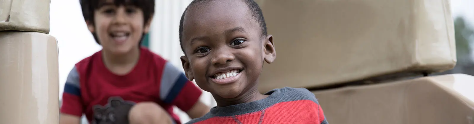 A smiling boy in a red and gray shirt sits in the foreground with another smiling boy in a red shirt behind him, both near stacked cardboard boxes.