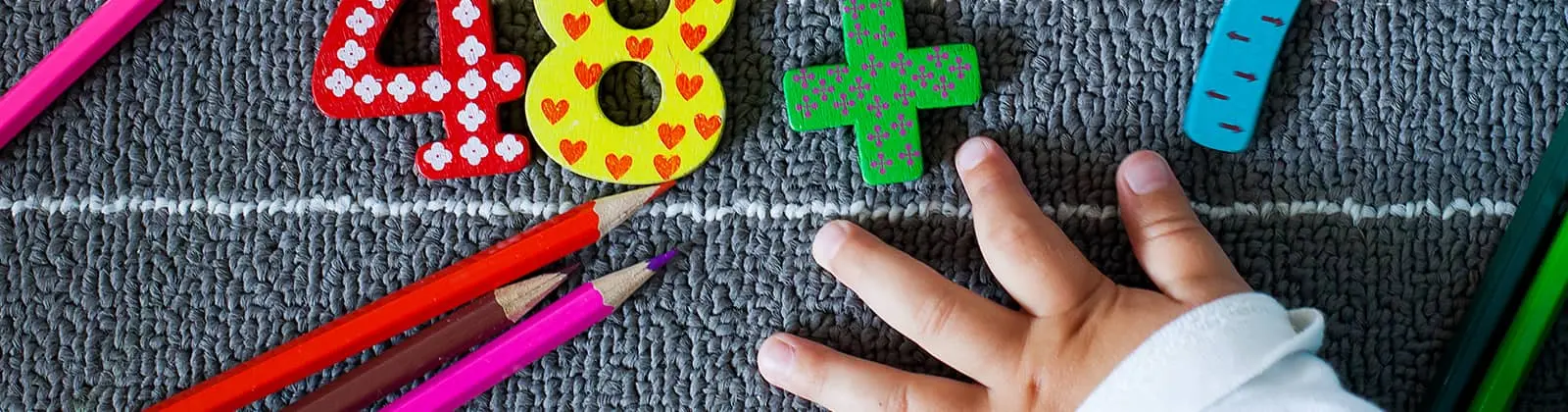 A child's hand rests on a textured gray surface beside colorful wooden numbers, a plus sign, and several colored pencils.