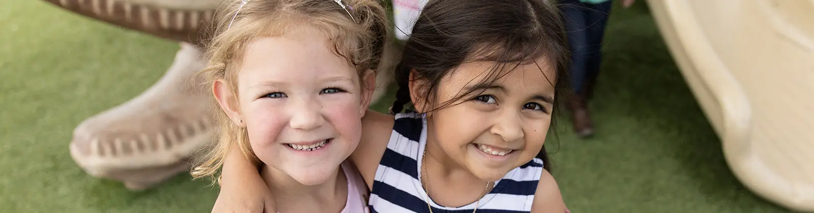 Two young girls smile at the camera while one girl rests her arm around the other's shoulder, with a large sculpted object and green ground visible behind them.