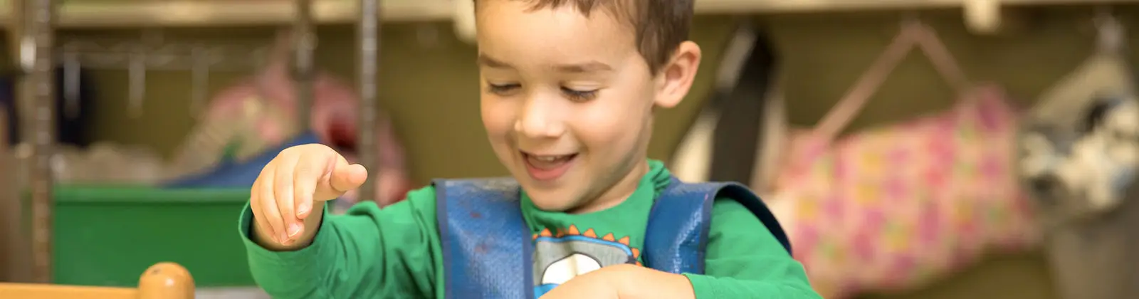 A young boy in a green shirt and blue vest smiles while playing with a wooden toy at a table.