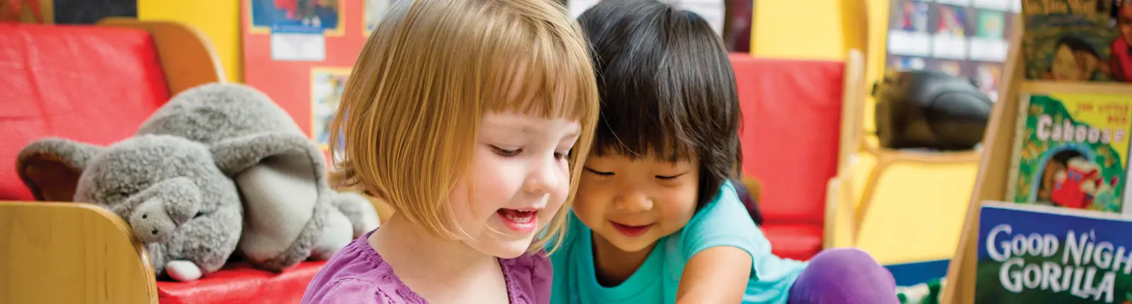 Two young girls sit closely together looking down at something out of frame, with a stuffed gray animal resting on a red chair behind them.