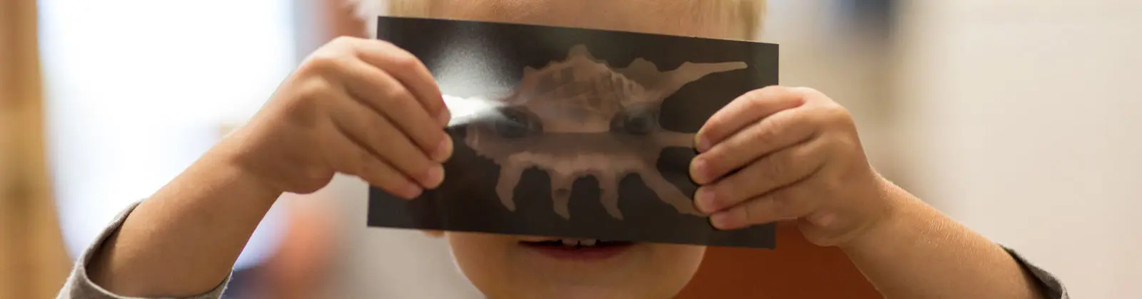 A child holds a dark card with a shell image in front of their face, covering their eyes.