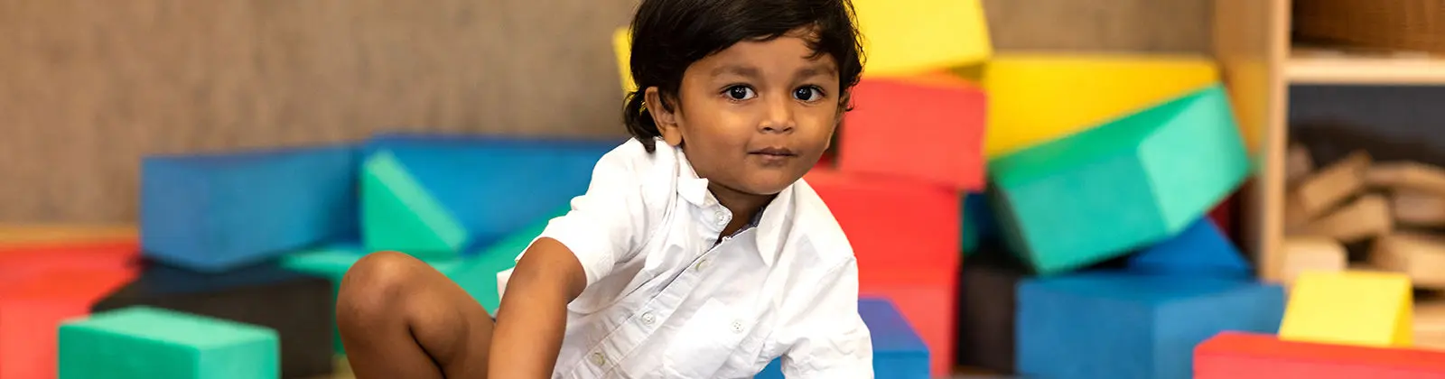 A young child in a white shirt sits among colorful foam blocks in a play area.