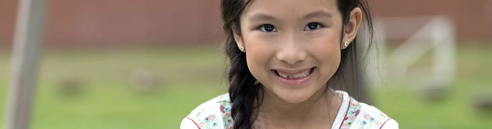 A smiling girl with a braided ponytail and stud earrings wears a white top with floral embroidery while standing outdoors.