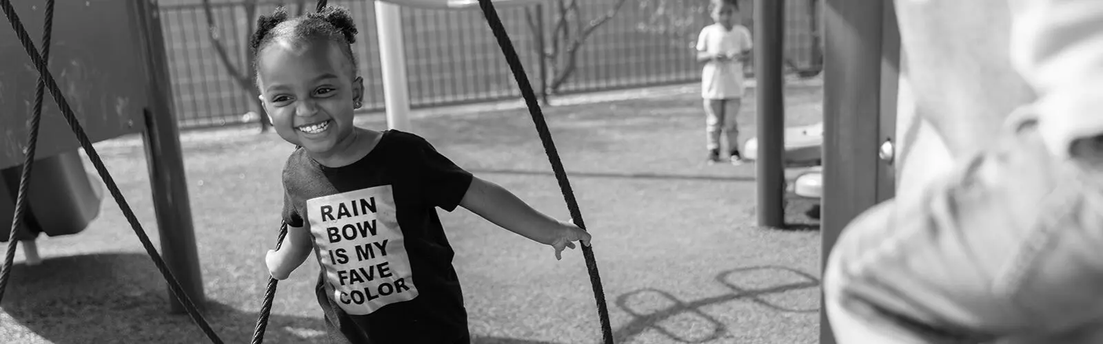 A smiling girl in a shirt reading 'RAIN BOW IS MY FAVE COLOR' holds playground ropes while another child stands near a fence in the background.