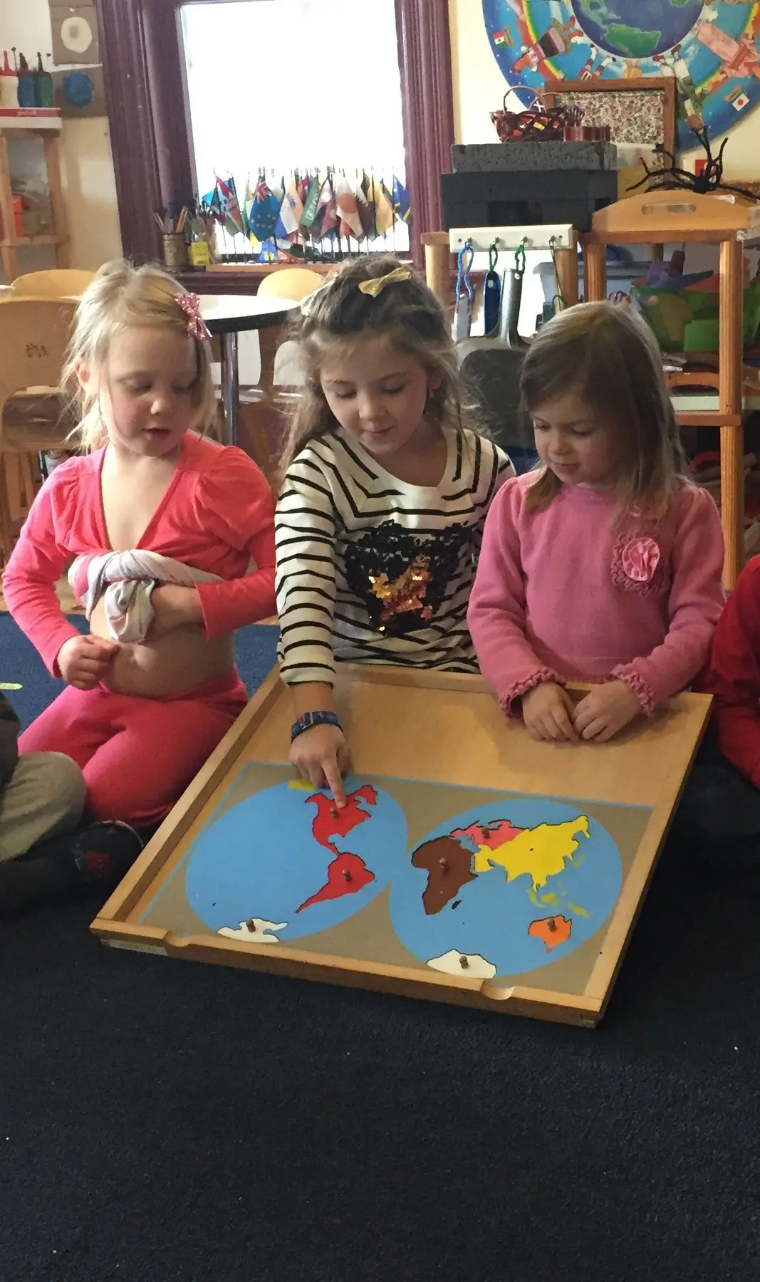 Three young girls sit on a dark carpet around a wooden puzzle map of the world, with one girl pointing at North America while another adjusts her shirt.