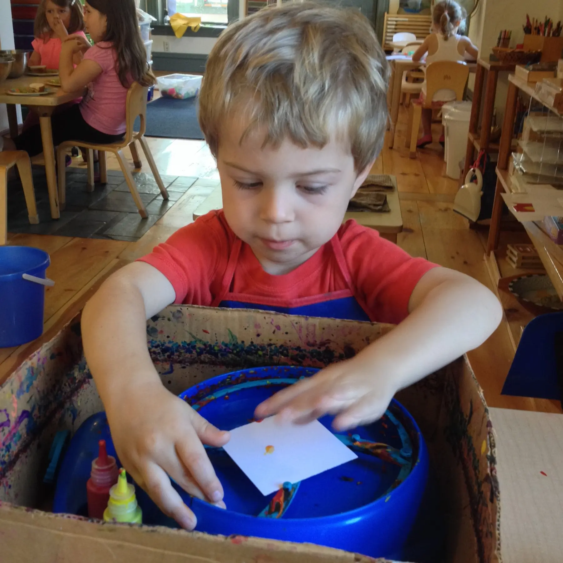 A young boy in a red shirt and blue apron presses a small white paper into a blue plastic tray inside a cardboard box while two girls sit at a table in the background.
