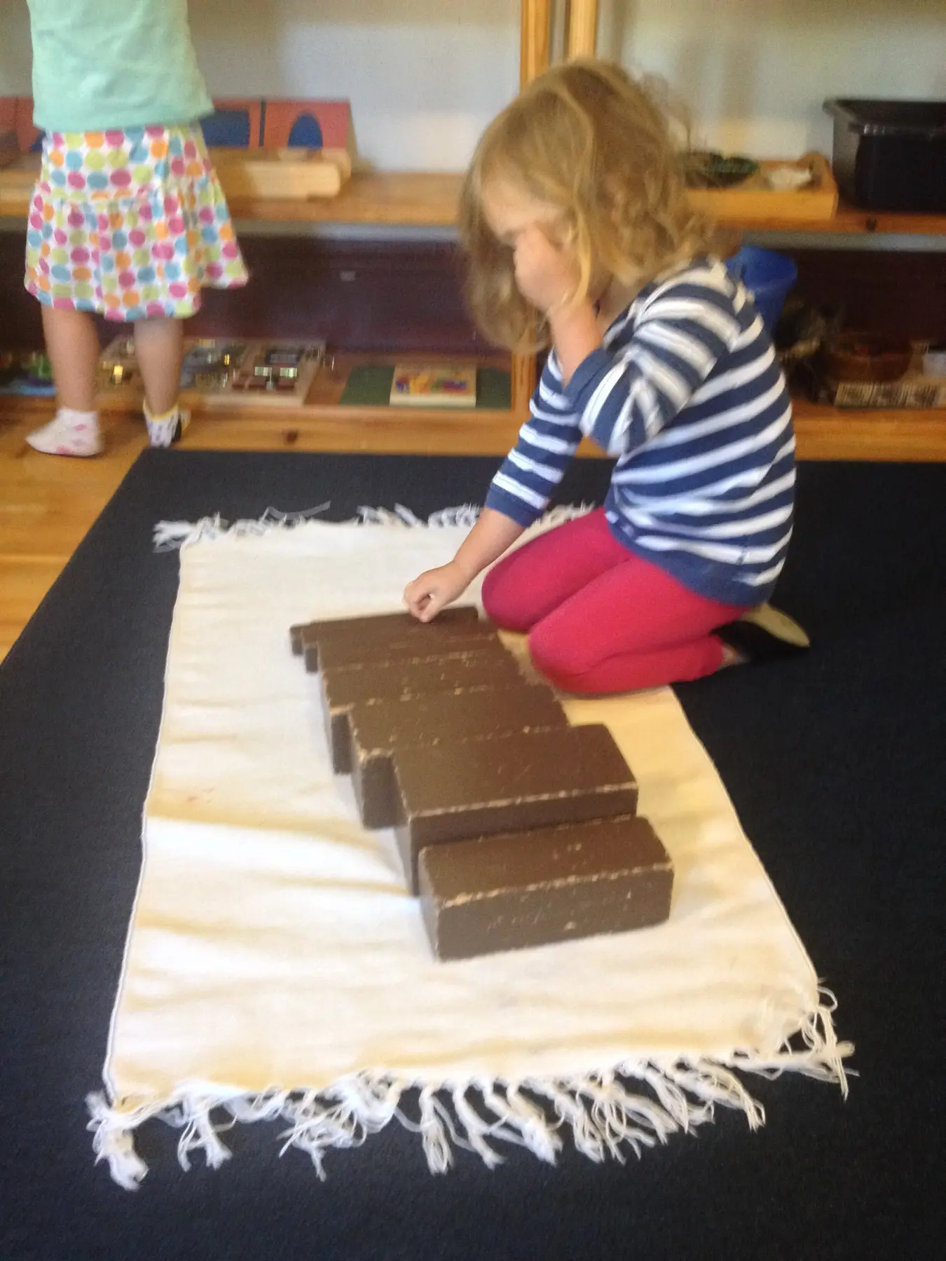A child in a striped shirt and pink pants kneels on a fringed mat arranging rectangular brown blocks while another child stands nearby.