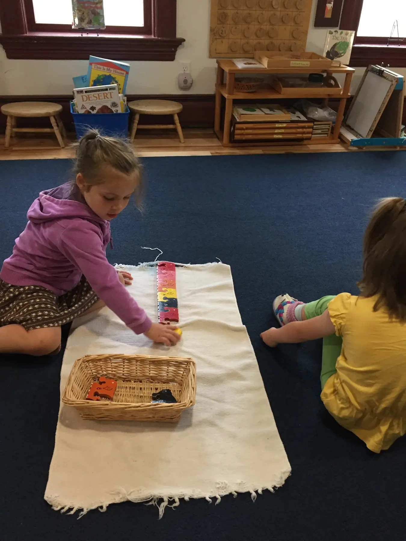 Two young girls sit on a blue carpet as one places a small block on a long segmented strip laid on a white mat beside a woven basket.