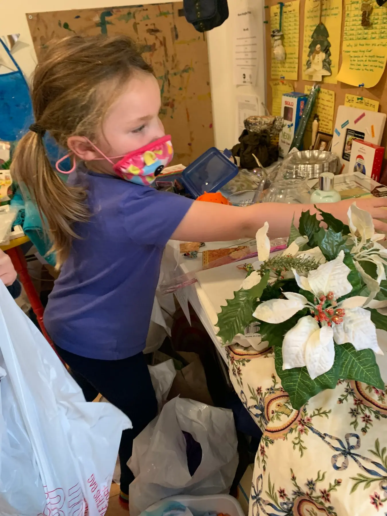 A young girl wearing a colorful face mask and blue shirt reaches toward a table holding a white poinsettia decoration in a cluttered room.