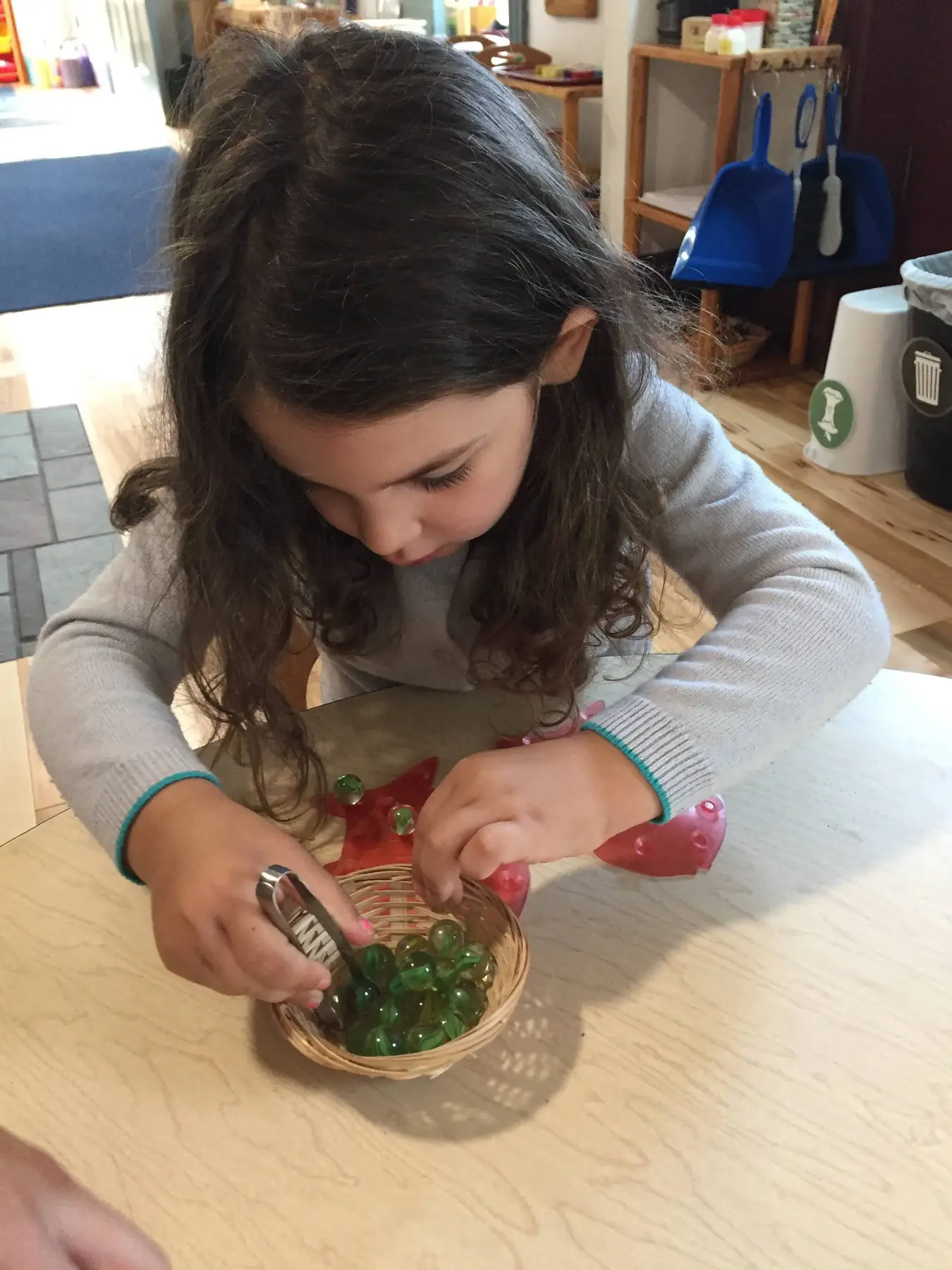 A young girl in a gray sweater uses tongs to move green glass stones into a small woven basket on a wooden table.