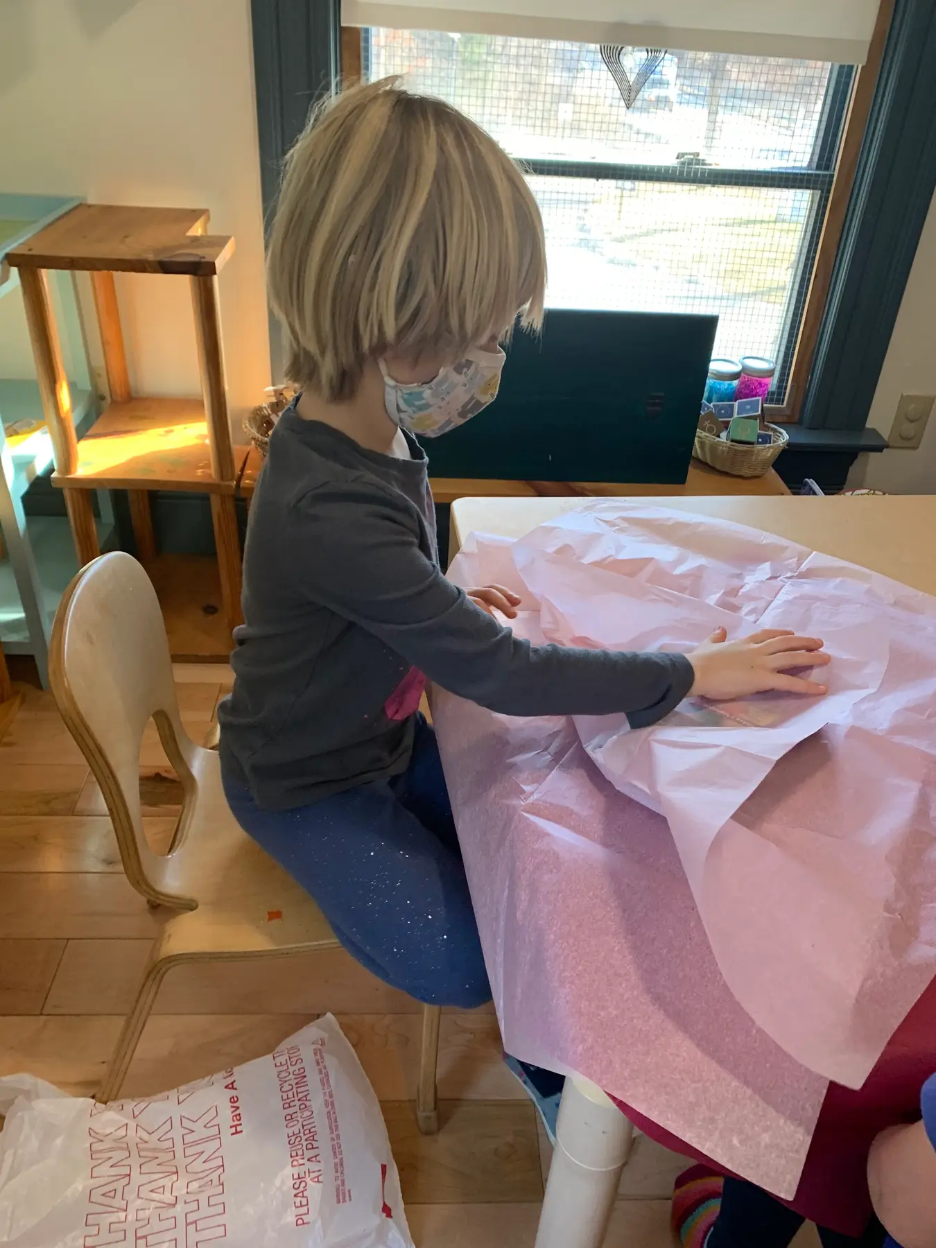 A child wearing a face mask sits at a table and presses hands onto pink tissue paper.