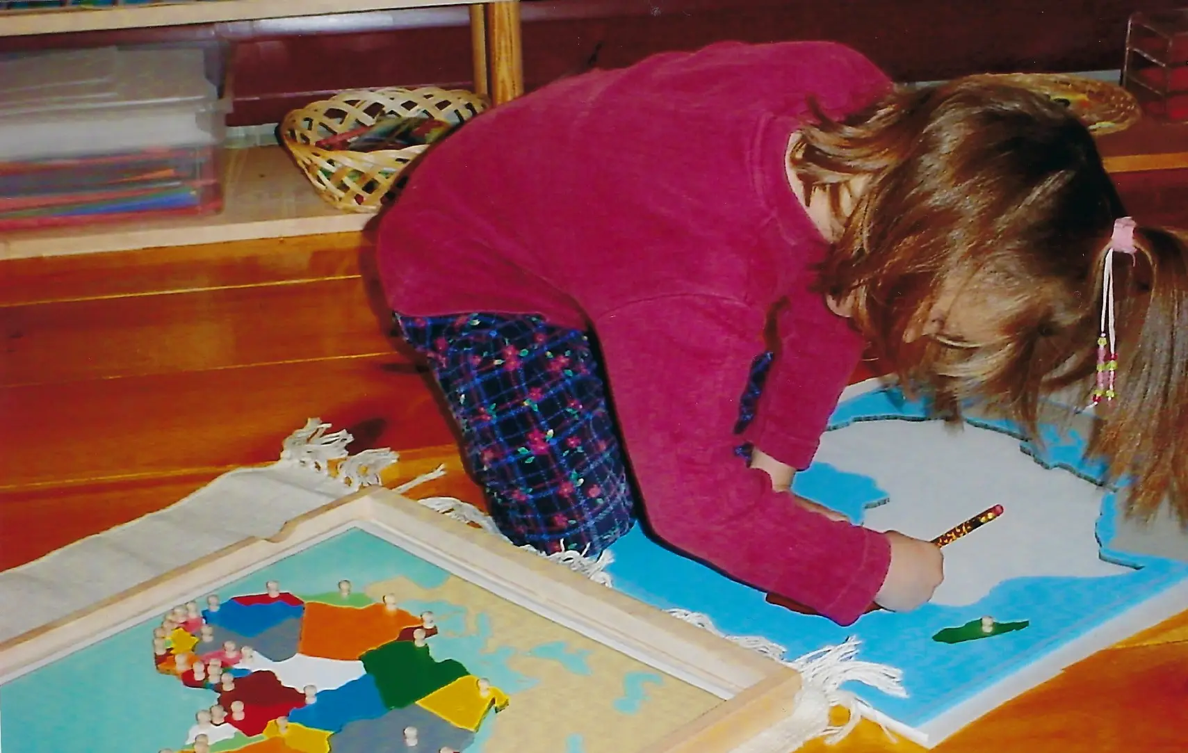 A child in a pink top and plaid pants leans over a framed map puzzle on a rug, holding a marker over a blue and white map.