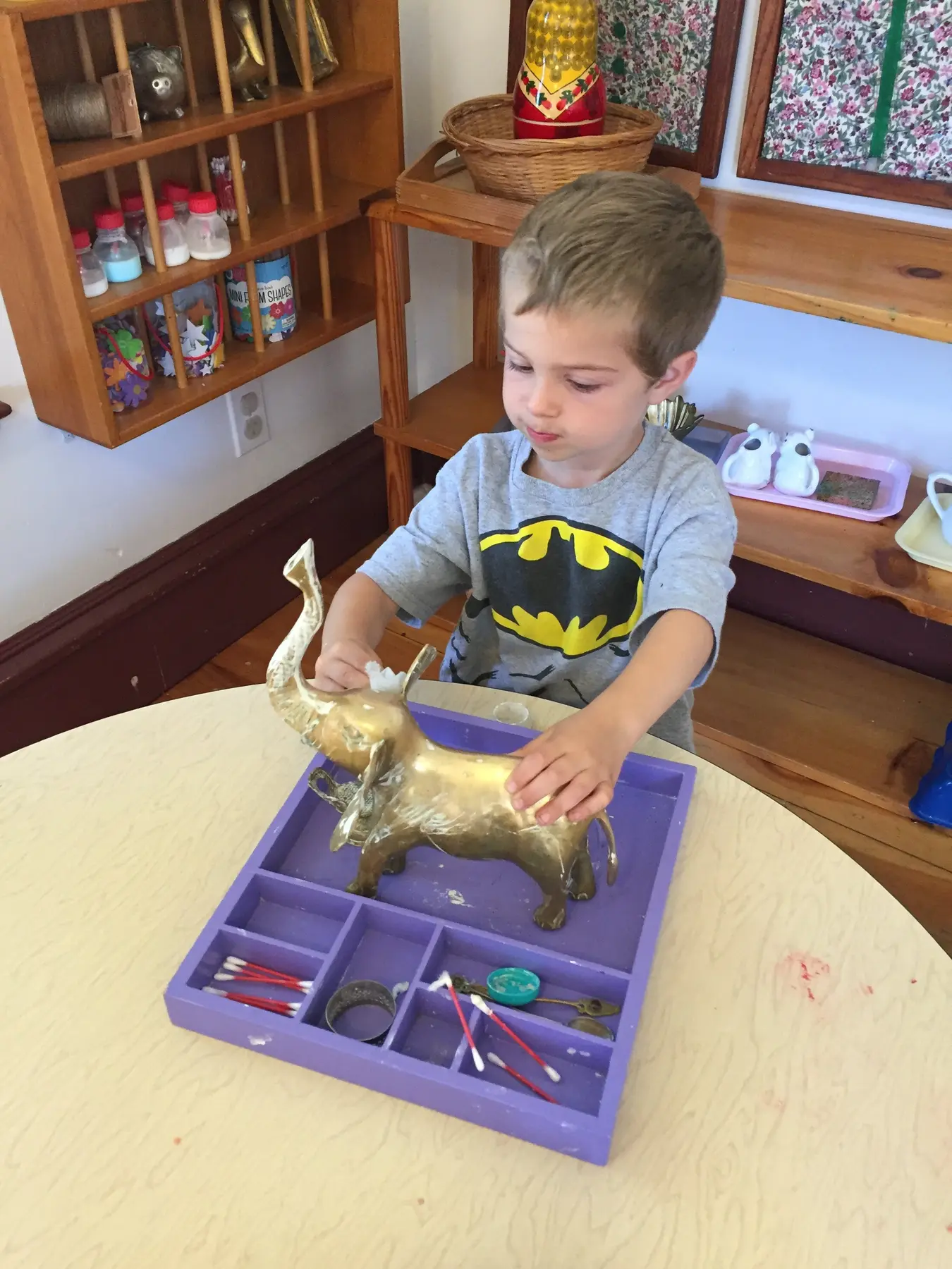 A young boy in a Batman shirt stands at a table and uses a cotton swab to clean a gold elephant figurine on a purple sorting tray.