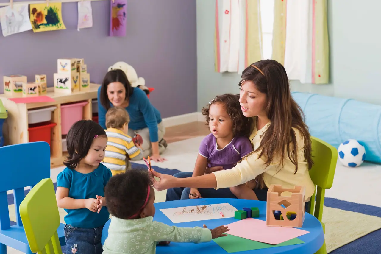 Two women sit with four young children around a blue table with paper and crayons, while a wooden shelf with blocks and a blue tunnel sit in the background.