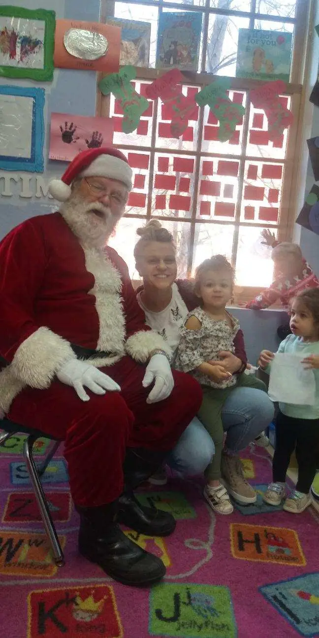 A person in a Santa costume sits on a chair beside an adult and three children in a room with a decorated window and a letter-patterned rug.