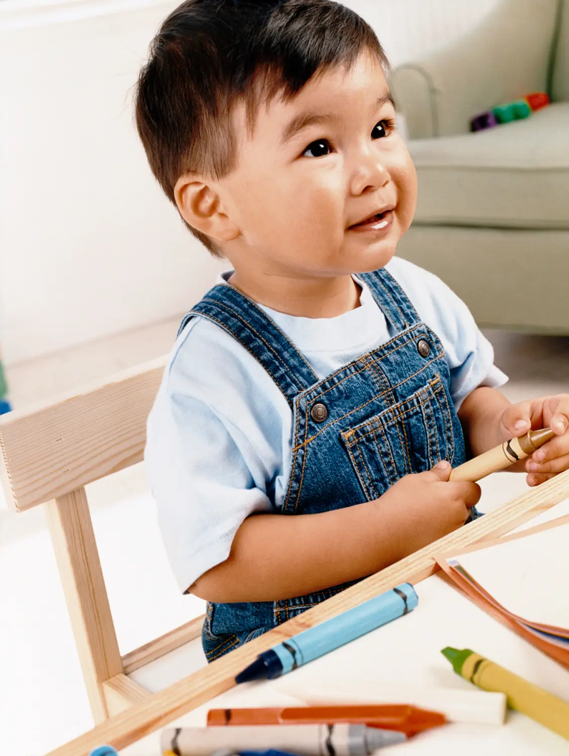 A young boy in denim overalls sits at a wooden desk holding a crayon, with several crayons and paper on the table.