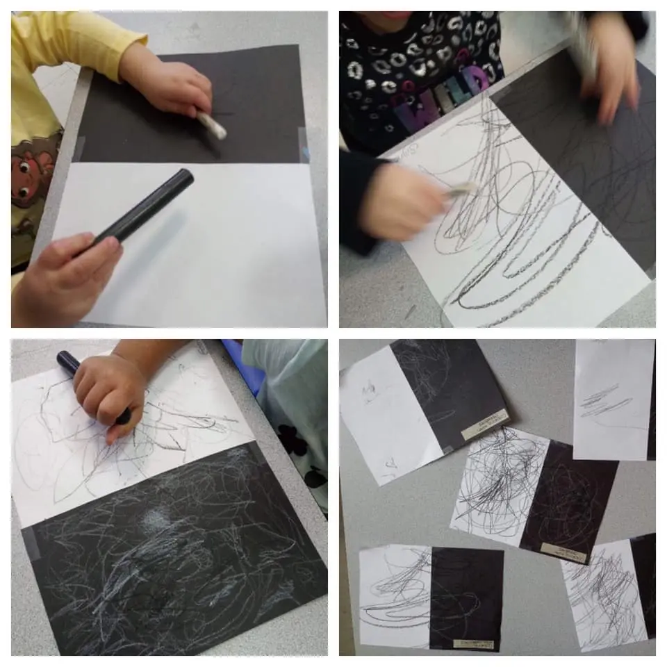 Children's hands use chalk and a black marker to draw scribbles on white paper and black construction paper taped to a table.