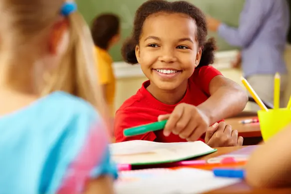 A smiling girl in a red shirt holds a green marker over paper at a table while other children and an adult are visible in the classroom.