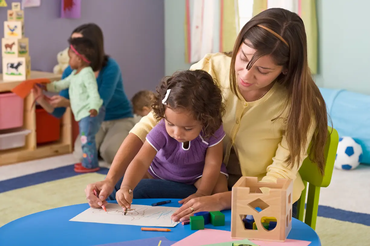 A woman in a yellow cardigan guides a toddler in a purple shirt as she draws on paper at a blue table, with other children and an adult in the background.