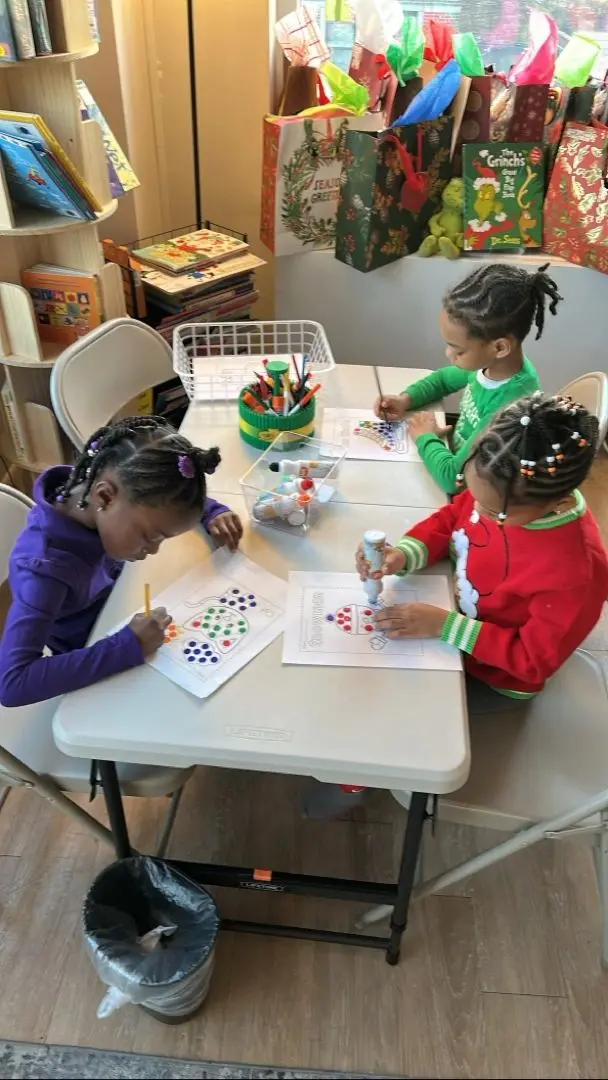 Three children sit at a white table coloring dot worksheets with markers and a paint dauber, surrounded by a supply caddy, a bin of materials, and gift bags in the background.