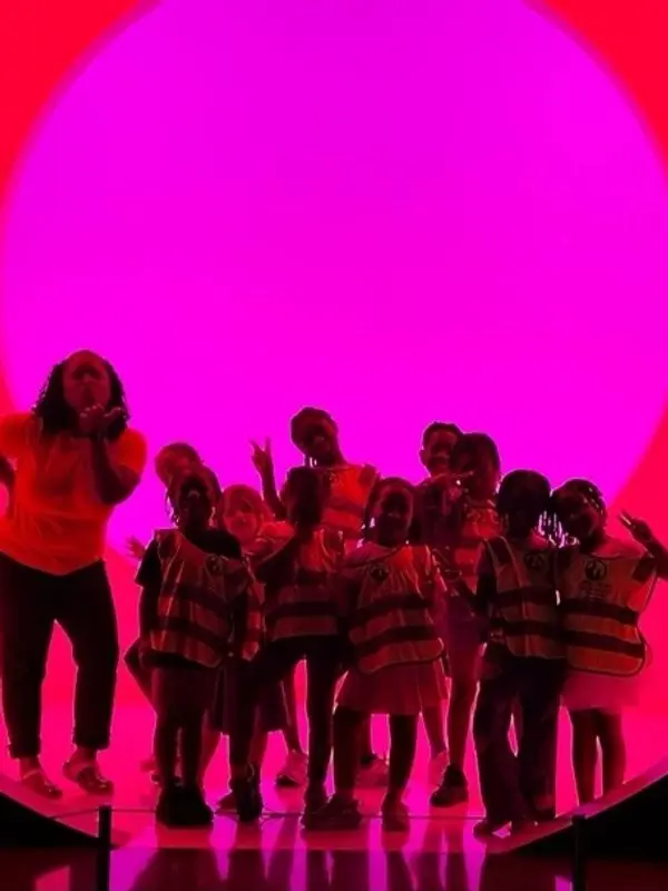 A group of children and one adult stand together in front of a large glowing pink circular light, with several children wearing matching safety vests.