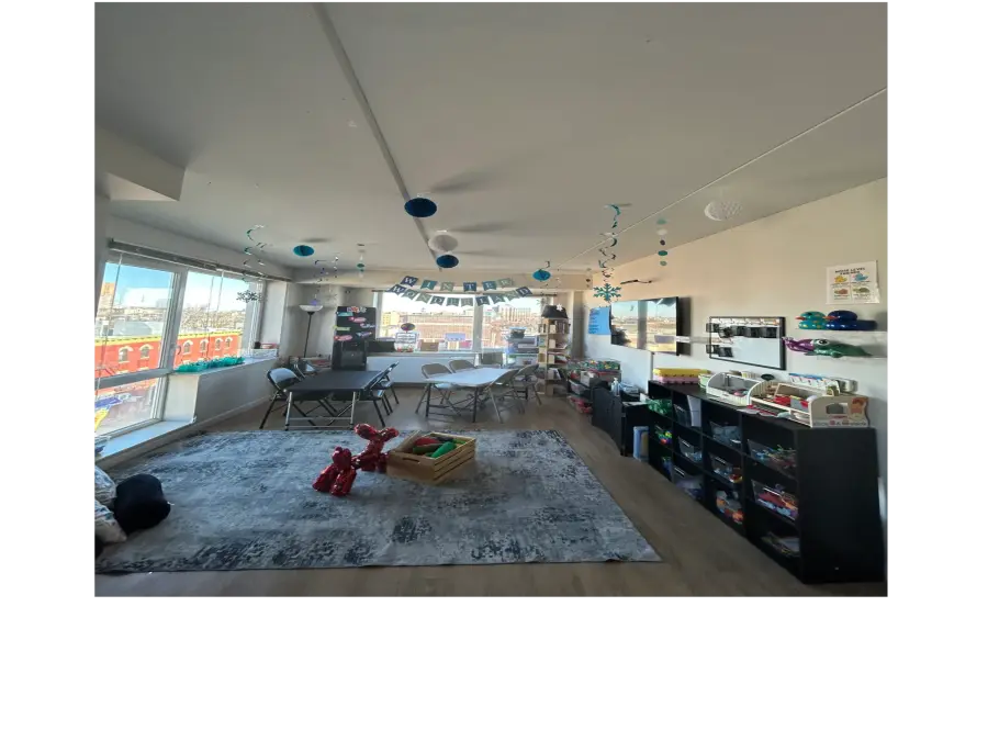 An empty classroom with a large patterned rug, a red stuffed animal, a wooden toy box, tables and chairs, and black shelving units filled with toys.