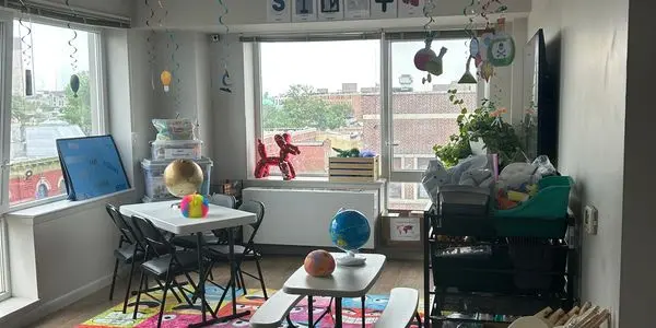 A sunlit classroom with small tables and chairs, a colorful rug, a globe, a red balloon dog, and storage shelves near large windows.