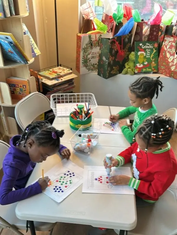 Three children sit at a table coloring dot worksheets with markers and crayons, with a supply caddy and bins nearby.