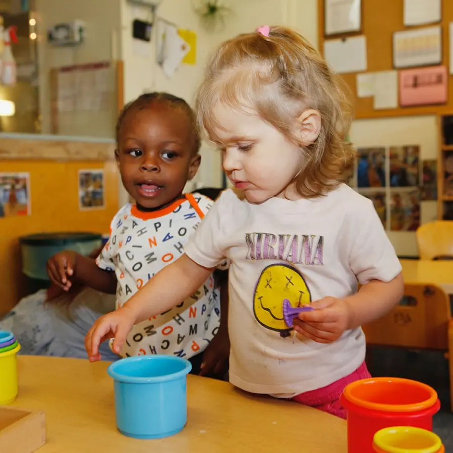 A toddler girl in a Nirvana shirt holds a purple toy while reaching toward a blue cup on a table, as a toddler boy in a patterned shirt watches beside her.