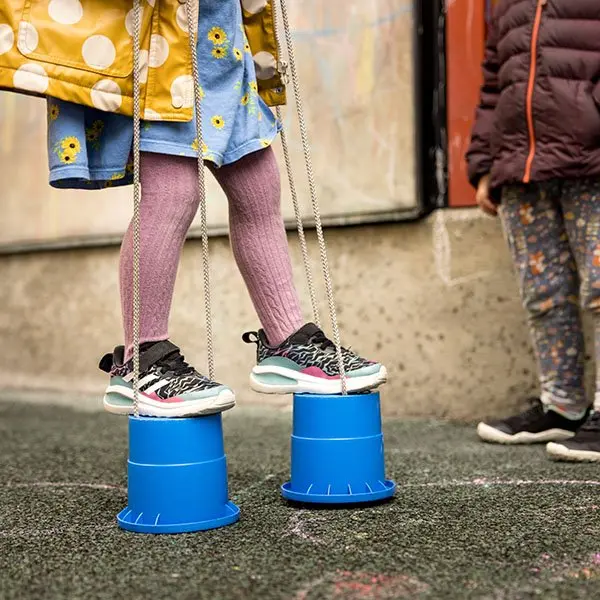 A child stands on two blue upside-down buckets with ropes attached while another child in a puffy jacket stands nearby on pavement.