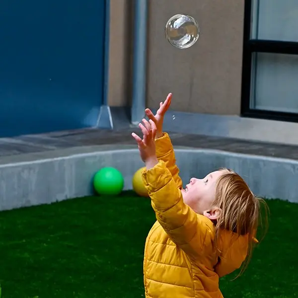 A child in a yellow puffer jacket reaches up toward a large soap bubble floating above them, with two balls resting on artificial grass behind.