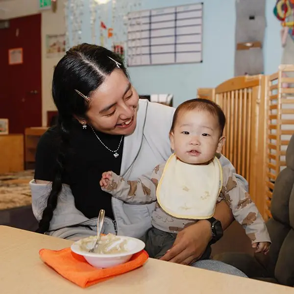 A person with braided hair and hair clips smiles while holding a baby in a bib seated at a table with a bowl of food and a spoon on an orange napkin.