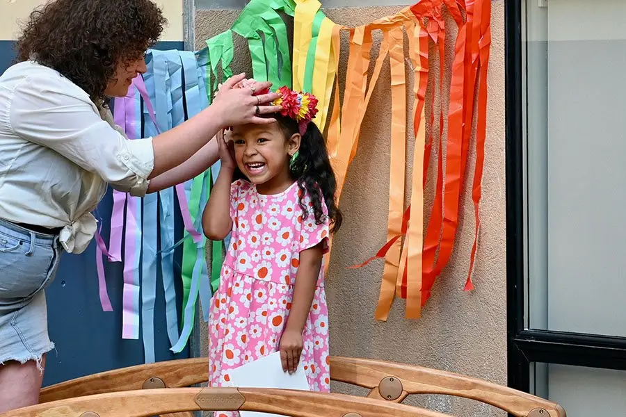 A woman adjusts a flower headband on a smiling girl in a pink floral dress as she holds a white paper, standing before colorful hanging paper streamers.