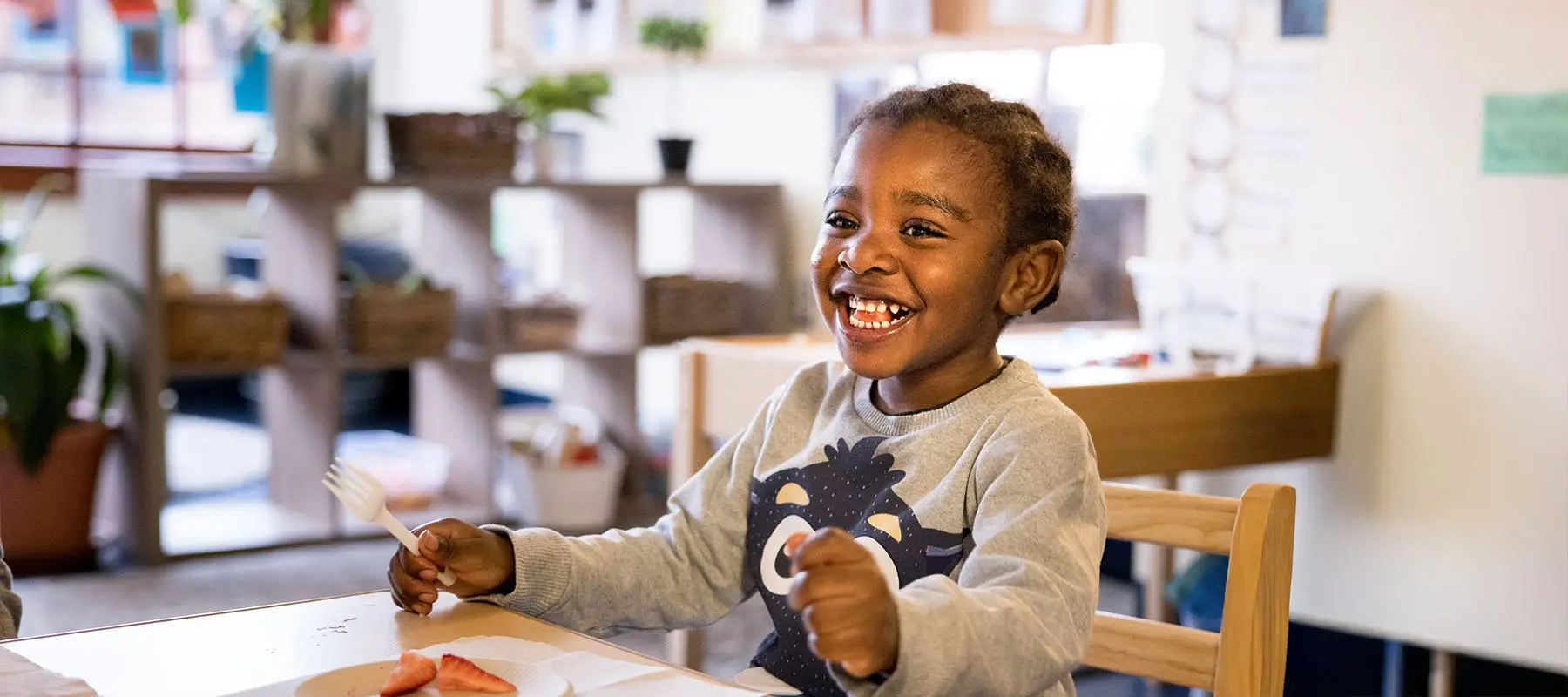 A smiling child sits at a table holding a white fork over a plate with sliced strawberries.