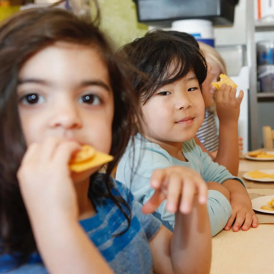 Three children sit at a table eating yellow snacks, with one child in the foreground holding a chip to their mouth and another reaching forward.